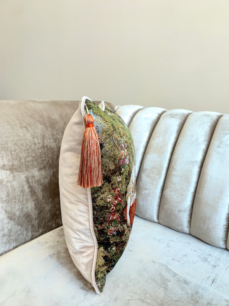 May include: A decorative throw pillow with a tapestry-like design featuring a floral pattern in shades of green, brown, and red. The pillow has a white velvet border and a long, orange tassel.