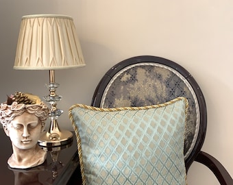 May include: A light blue throw pillow with a geometric pattern and gold trim sits on a dark brown chair with a patterned fabric. The chair is in front of a white wall. A table lamp with a cream-colored shade is on a dark brown table to the left of the chair.
