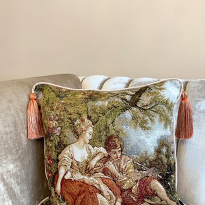 May include: A decorative throw pillow with a tapestry design featuring a couple in a garden setting. The pillow has a green, brown, and beige color scheme with orange tassels on the corners.