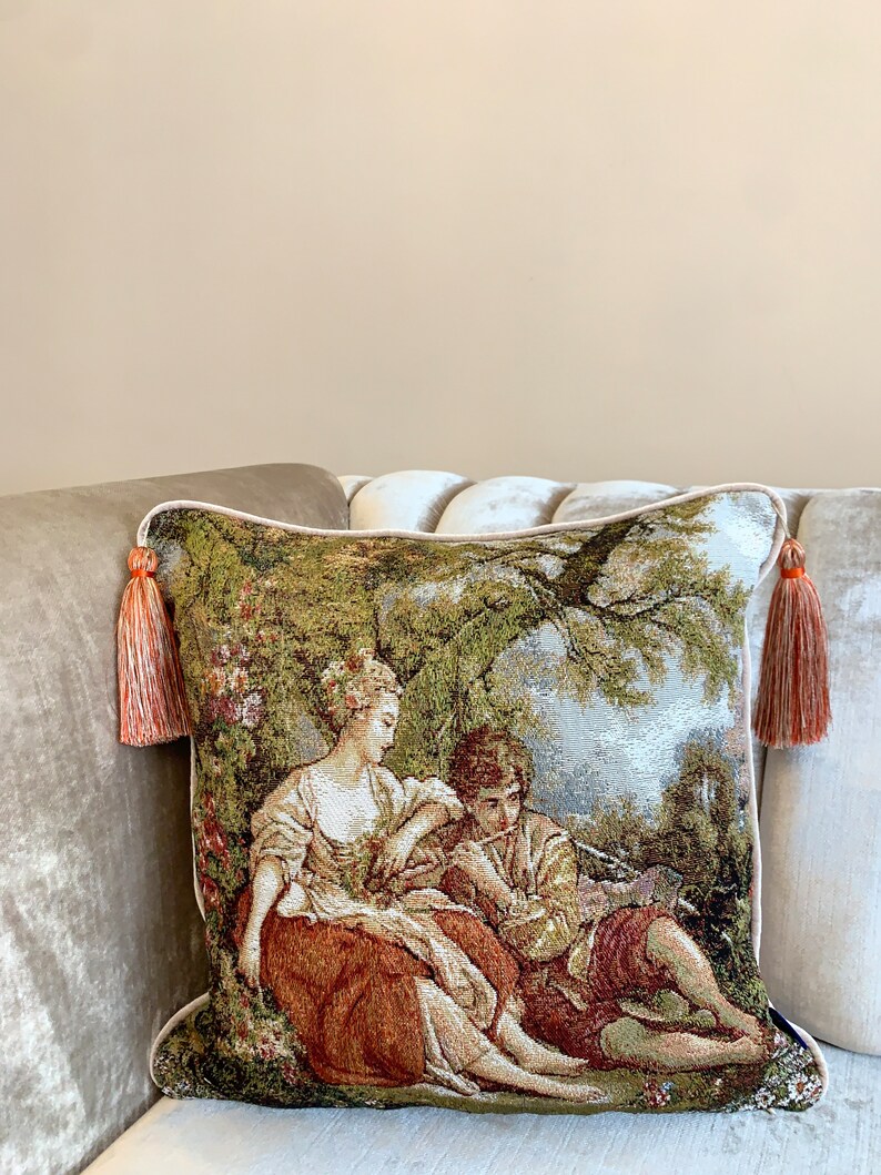 May include: A decorative throw pillow with a tapestry design featuring a couple in a garden setting. The pillow has a green, brown, and beige color scheme with orange tassels on the corners.