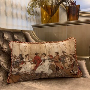 May include: A decorative cushion with a tapestry design featuring a group of people dancing in a ballroom. The cushion has a red and gold trim.