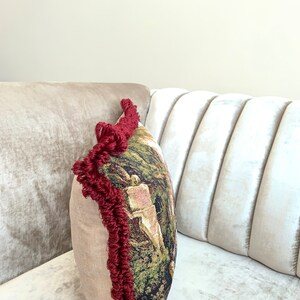 Op de afbeelding: Een decoratief kussen met een bordeauxrode franje rand en een tapijt-achtig ontwerp met een bos sc&egrave;ne. Het kussen ligt op een licht beige bank.
