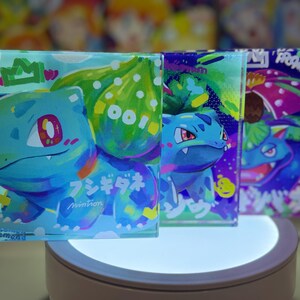 May include: Three square glass coasters featuring a cartoon illustration of a blue and green Pokemon character. The coasters are on a white circular platform with a light shining from below. The text on the coasters reads "Pokemon" and "Miniman".