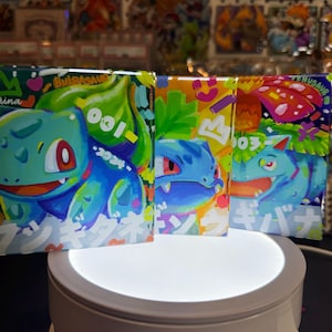 May include: Three colorful square cards featuring a cartoon illustration of a blue and green Pokemon character. The cards have a white background with colorful accents and Japanese text. The cards are on a white rotating display stand.