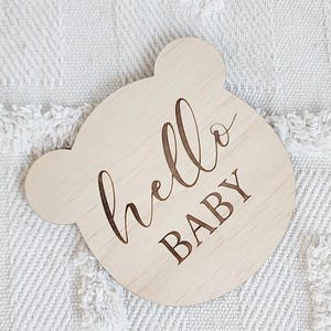 May include: A wooden sign shaped like a bear's head with the text "hello baby" engraved on it.