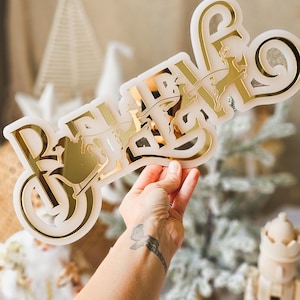 May include: A gold-coloured "Believe" sign with a silhouette of Santa Claus and reindeer in a sleigh. The sign is held in a hand with a tattoo on the wrist.