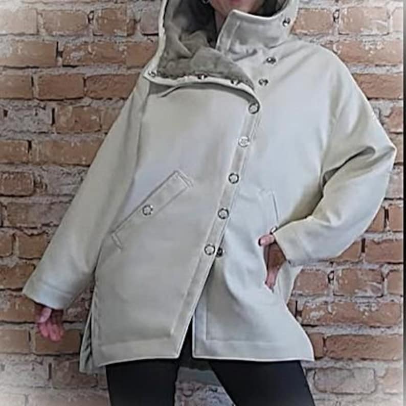 May include: A light beige hooded jacket with a high collar and a series of silver snap closures. The jacket has a diagonal closure and a pocket on the left side. The interior of the hood appears to be a soft, textured material.