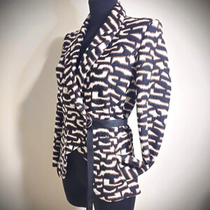 May include: A patterned blazer in navy, white, and brown, displayed on a mannequin. The blazer features a unique geometric design and is cinched at the waist with a black belt. The jacket has a notched collar and long sleeves.