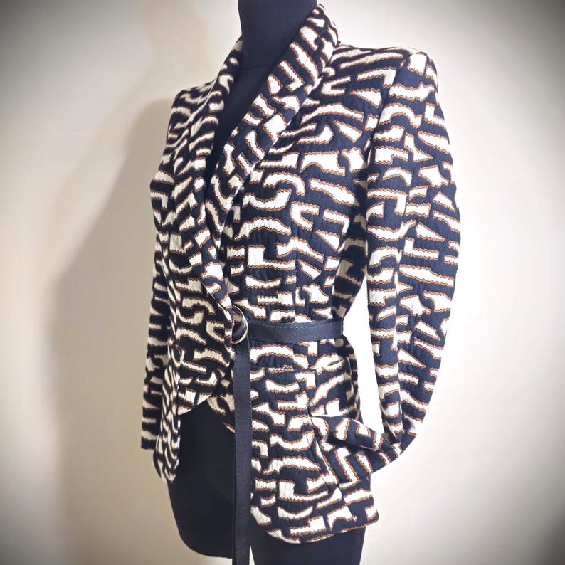 May include: A patterned blazer in navy, white, and brown, displayed on a mannequin. The blazer features a unique geometric design and is cinched at the waist with a black belt. The jacket has a notched collar and long sleeves.