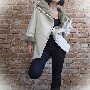 May include: A cream-colored coat with a faux fur-lined hood and cuffs. The coat has a button-up front and a relaxed fit. The model is wearing black pants and a black top. The background is a brick wall.