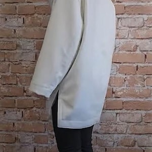 May include: A light-colored, hooded jacket with a longline silhouette. The jacket features a high collar and a front zipper closure. The model is wearing black pants and white sneakers. The background is a brick wall.