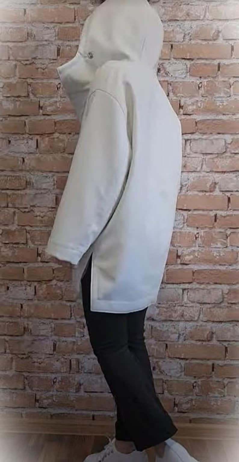 May include: A light-colored, hooded jacket with a longline silhouette. The jacket features a high collar and a front zipper closure. The model is wearing black pants and white sneakers. The background is a brick wall.