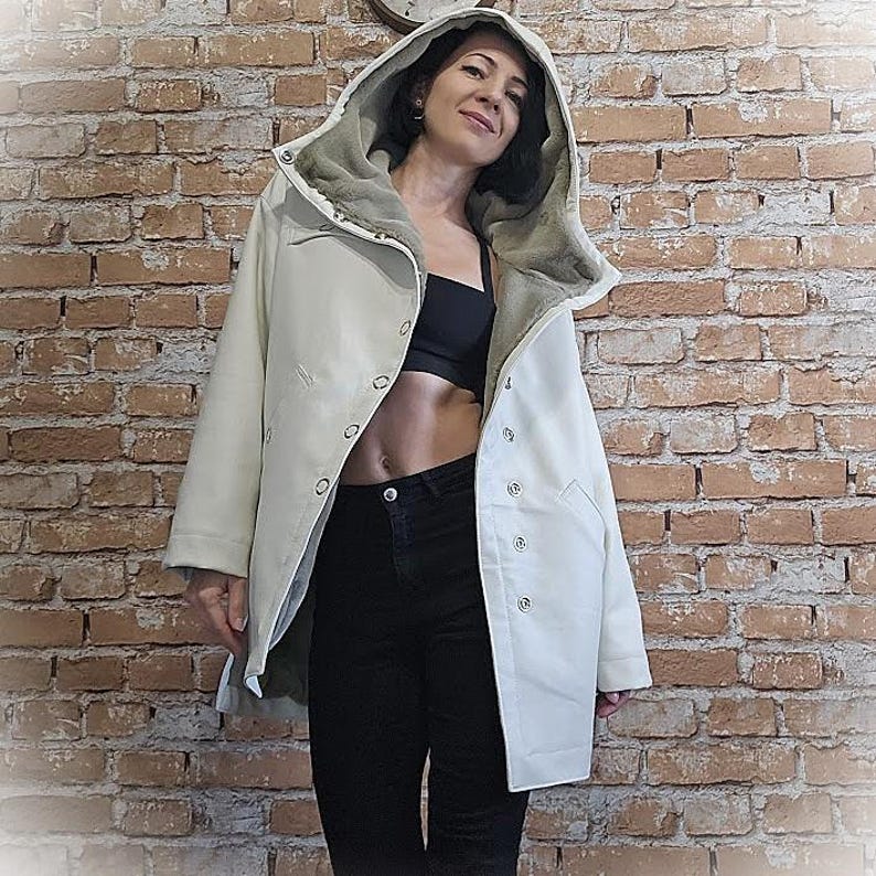 May include: Off-white hooded coat with a light green faux fur lining. The coat has a button-up front and two pockets. The person in the image is wearing a black top and black pants. The background is a brick wall.