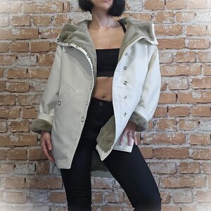 May include: A cream-colored coat with a faux fur-lined hood and cuffs. The coat has a button-up front and a relaxed fit. The model is wearing black pants and a black top. The background is a brick wall.