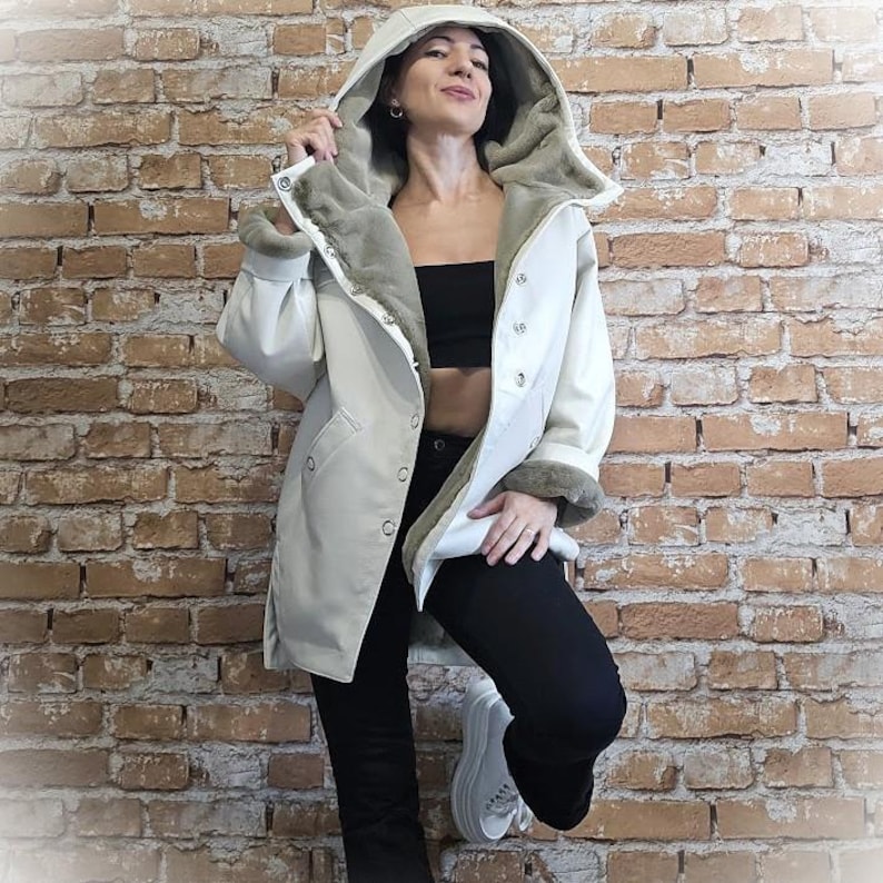 May include: A cream-colored hooded coat with a faux fur lining and trim. The coat features a button-up front and is worn with black pants and white sneakers. The coat's hood is up, and the lining is a soft gray color.