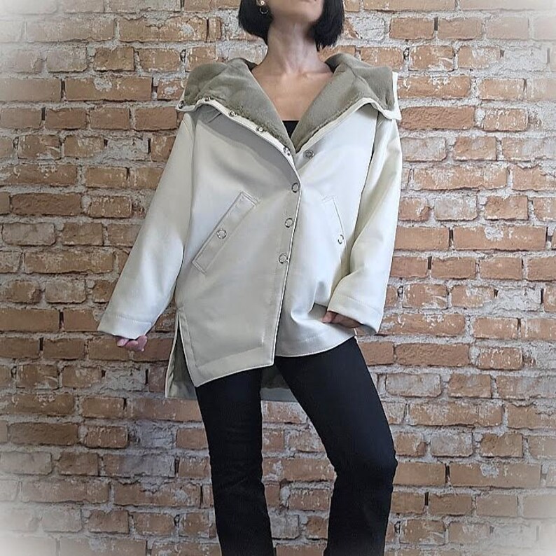 May include: Cream-colored hooded jacket with a light green faux fur lining. The jacket has a button-up front, side pockets, and a split hem. The model is wearing black pants.