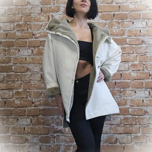 May include: Cream-colored coat with a faux fur-lined hood and cuffs. The coat features a button-up front and a relaxed fit. The lining of the hood and cuffs is a muted green color. The model is wearing black pants and a black top.