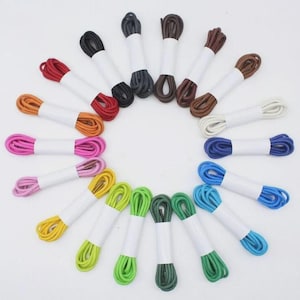 Könnte beinhalten: Eine Sammlung farbenfroher Schnürsenkel in verschiedenen Farbtönen, darunter Rot, Orange, Rosa, Gelb, Grün, Blau und Braun. Jedes Paar ist ordentlich mit einem weißen Papierband gebündelt. Die Schnürsenkel sind kreisförmig angeordnet.