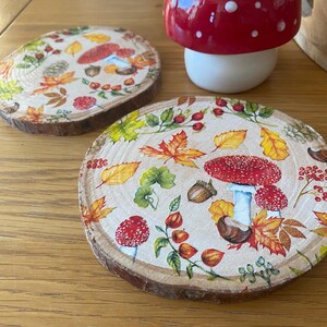May include: Two round wooden coasters featuring a printed autumn design of red mushrooms, brown acorns, and colourful fall leaves in shades of yellow, orange, and green. The coasters have a natural wood grain edge and a smooth, flat top surface.