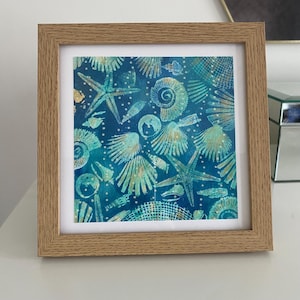 May include: Square framed artwork with a blue backdrop, showcasing a pattern of seashells and starfish in teal and gold hues. The artwork is enclosed in a light brown wooden frame, creating a coastal theme.