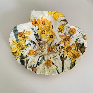 May include: A decorative seashell-shaped trinket dish with a floral design featuring yellow daffodils and white flowers. The dish holds three pairs of gold hoop earrings. The background is white.