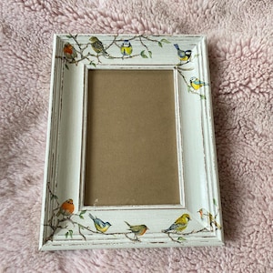 May include: White picture frame with a distressed finish and a floral design featuring colorful birds. The frame has a white mat and a clear glass front.