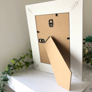 May include: White picture frame with a brown cardboard backing and a kickstand. The frame has a decorative design on the edges.