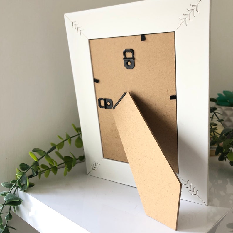 May include: White picture frame with a brown cardboard backing and a kickstand. The frame has a decorative design on the edges.