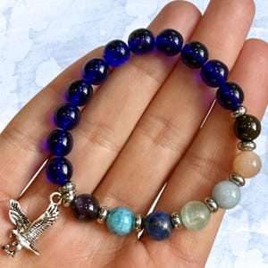 Puede incluir: Una pulsera elástica con el texto "EAGLE WISDOM CHAKRA STRETCH BRACELET". La pulsera presenta cuentas azul oscuro, varias cuentas de piedras preciosas de colores y un dije de águila plateado. La pulsera se sostiene en una mano.