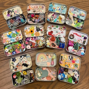 Può includere: Una collezione di piccole scatole di metallo rettangolari, ciascuna riempita con un assortimento di bigiotteria, ciondoli e oggetti decorativi colorati. Alcune scatole presentano decorazioni a tema festivo, mentre altre hanno una varietà di perline, bottoni e figurine in miniatura. Il testo "Feel Your Feelings" è visibile su una scatola.