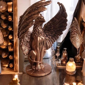 Peut inclure: Statue de griffon en bronze, avec des ailes déployées et des détails de plumes. La créature mythique est assise sur une base circulaire. Objet décoratif avec une finition bronze.