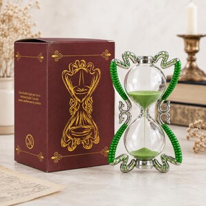 Può includere: Una clessidra con sabbia verde e una cornice color argento con accenti verdi a forma di serpente. Accanto si trova una scatola rettangolare marrone scuro decorata con disegni a linee dorate di una clessidra e del testo piccolo sul lato.