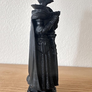 May include: A black chess piece depicting a knight in full armor, including a helmet with a visor and a long cape. The piece is standing on a hexagonal base and is made of a solid, dark material.