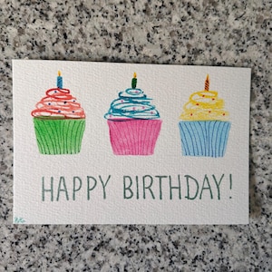 Può includere: Un biglietto d'auguri rettangolare bianco con tre cupcake disegnati a mano con glassa colorata e candeline accese. Il primo cupcake ha la glassa verde e una candela blu, il secondo rosa con glassa blu, il terzo blu con glassa gialla. Il testo HAPPY BIRTHDAY! è stampato in verde.