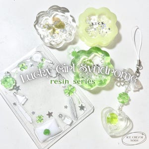 May include: A collection of resin art pieces with a lucky theme. Includes clear and green flower-shaped charms, a phone case, and a heart-shaped charm with a strap. The text "Lucky Girl Syndrome" and "resin series" are visible.