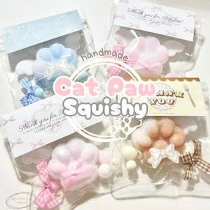 Puede incluir: Tres bolsas de plástico transparente con juguetes blandos con forma de pata de gato hechos a mano. Cada bolsa tiene un juguete blando de un color diferente: azul, rosa y melocotón. Cada bolsa incluye una pequeña tarjeta de agradecimiento con el texto "¡Gracias por tu apoyo!" y "¡Gracias por tu apoyo! ¡Un día bonito comienza con cada postre! ICECREAM SODA STUDIO".