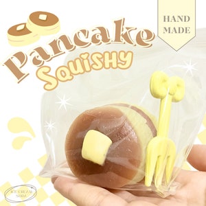 Può includere: Un giocattolo squishy a forma di pancake marrone e giallo in una busta di plastica trasparente con una forchetta di plastica gialla. La busta riporta il testo "Pancake Squishy" e "Hand Made".