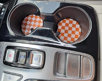 May include: Two orange and white checkered car coasters are placed inside the cup holders of a car's center console. The console is black and silver, with buttons for gear selection and parking.
