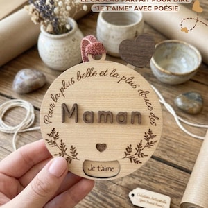 Peut inclure: Un disque en bois avec le mot "Maman" en lettres en relief, entouré de motifs floraux et de la phrase "Je t'aime". Le disque est tenu par une main, avec d'autres objets décoratifs en arrière-plan.