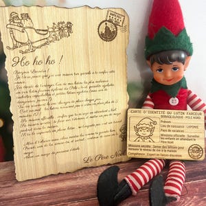 Peut inclure: Une lettre en bois du Père Noël avec les mots "Ho ho ho !" et un dessin du traîneau du Père Noël. À côté, une petite poupée elfe vêtue d'une tenue rouge et verte, tenant une carte d'identité.