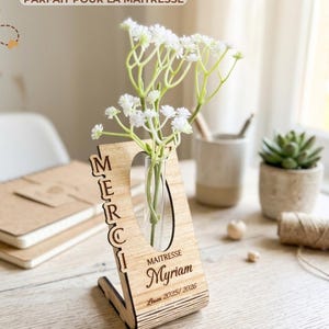 Peut inclure: Un vase en bois avec un tube en verre transparent contenant des fleurs blanches. Le vase a une découpe en forme de cœur et le mot « MERCI » verticalement sur le côté gauche. Le nom « Myriam » et le texte « Louen 2025/2026 » sont également visibles.