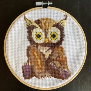 May include: A brown and yellow owl cross-stitch embroidery in a wooden hoop. The owl has large yellow eyes and is sitting on a white background.