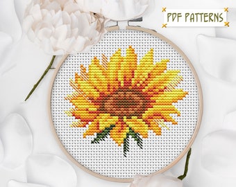 May include: A cross-stitch pattern of a sunflower with yellow petals and a brown center. The pattern is displayed in a wooden embroidery hoop.