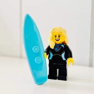 May include: A LEGO minifigure with blonde hair and a smiling expression, wearing a black wetsuit with light blue accents and a circular wave emblem. The figure stands next to a bright turquoise surfboard featuring two circular studs and the LEGO logo.
