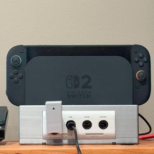 May include: A black Nintendo Switch console with blue and orange controllers is placed on top of a silver GameCube adapter. The adapter has several ports and a white adapter plugged in. The text "Nintendo Switch" is visible.
