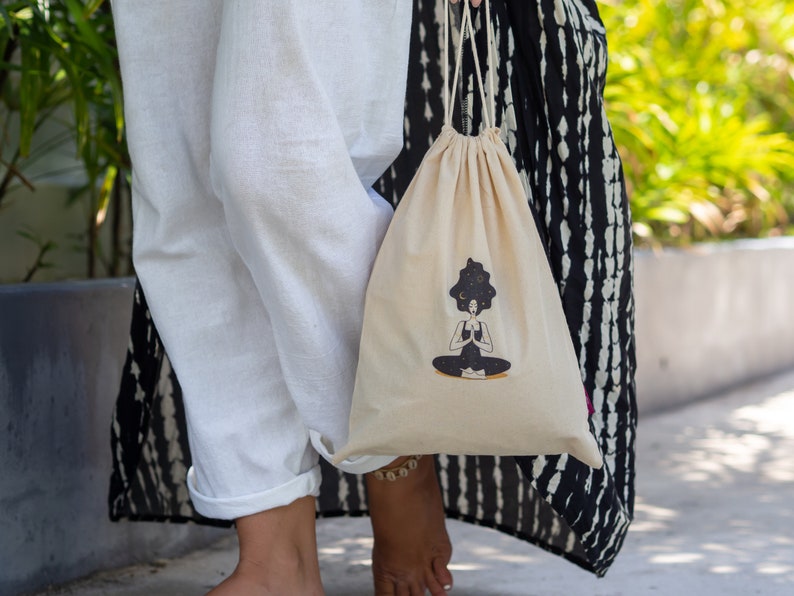 Puede incluir: Una bolsa de cord&oacute;n blanco con una ilustraci&oacute;n en blanco y negro de una persona meditando con un cielo estrellado sobre su cabeza. La bolsa est&aacute; hecha de algod&oacute;n natural y tiene un dise&ntilde;o simple.