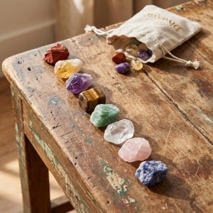 Op de afbeelding: Een verzameling van zeven kleurrijke chakra stenen gerangschikt op een rustiek houten oppervlak. De stenen zijn rood, geel, paars, bruin, groen, helder, roze en blauw. Een kleine beige zak met trekkoord en gouden tekst is open.