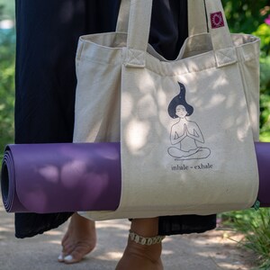 Puede incluir: Una persona llevando una bolsa de lona beige con una esterilla de yoga. La bolsa de lona tiene una ilustraci&oacute;n en blanco y negro de una persona meditando con el texto "inhale - exhale".