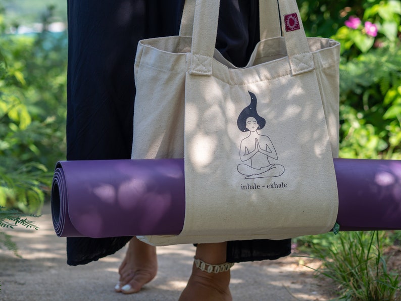 Puede incluir: Una persona llevando una bolsa de lona beige con una esterilla de yoga. La bolsa de lona tiene una ilustraci&oacute;n en blanco y negro de una persona meditando con el texto "inhale - exhale".