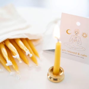 Pode incluir: Uma vela amarela acesa em um suporte dourado, com um saco de velas amarelas apagadas. Um cartão com um desenho de uma pessoa meditando e o texto "I am relaxed & calm" também é visível.